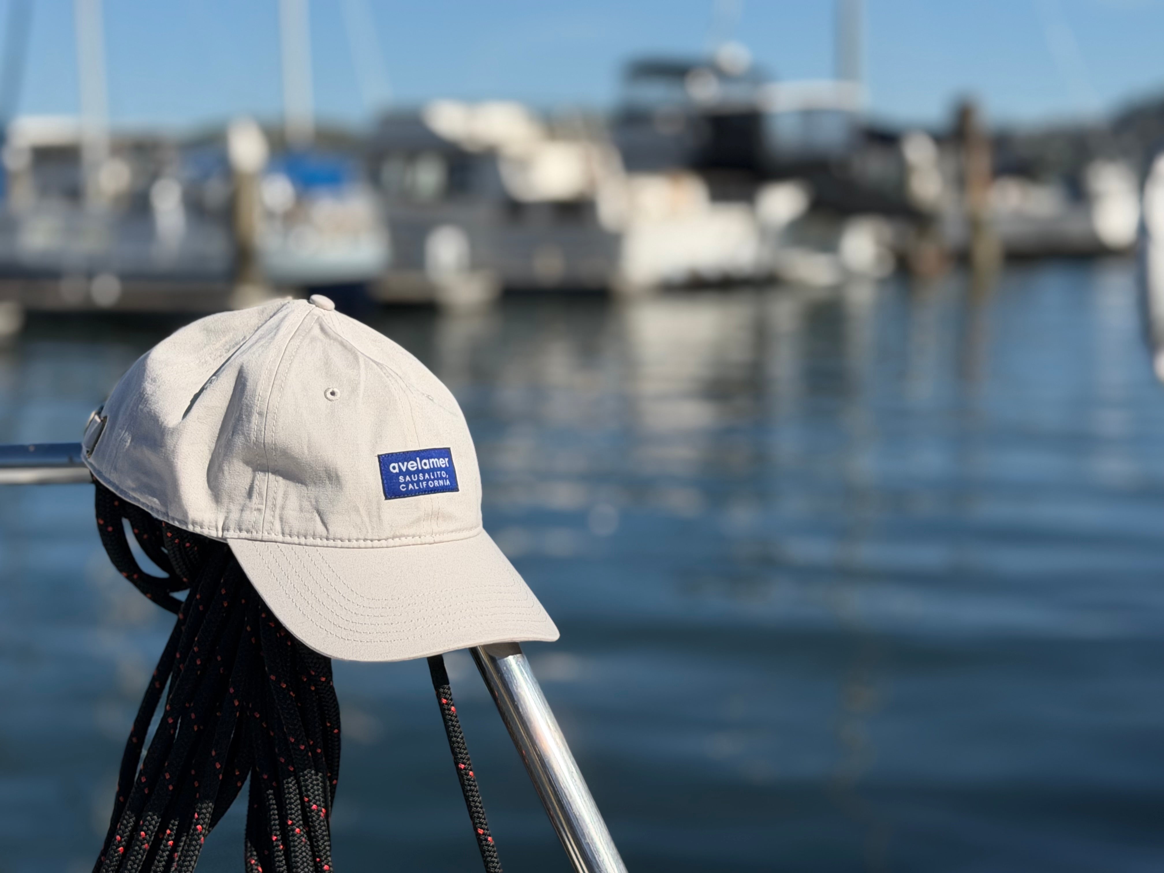 Avelamer Bridgeway Cap in Mainsail off-white with a blue woven brand label on a boat railing at a marina