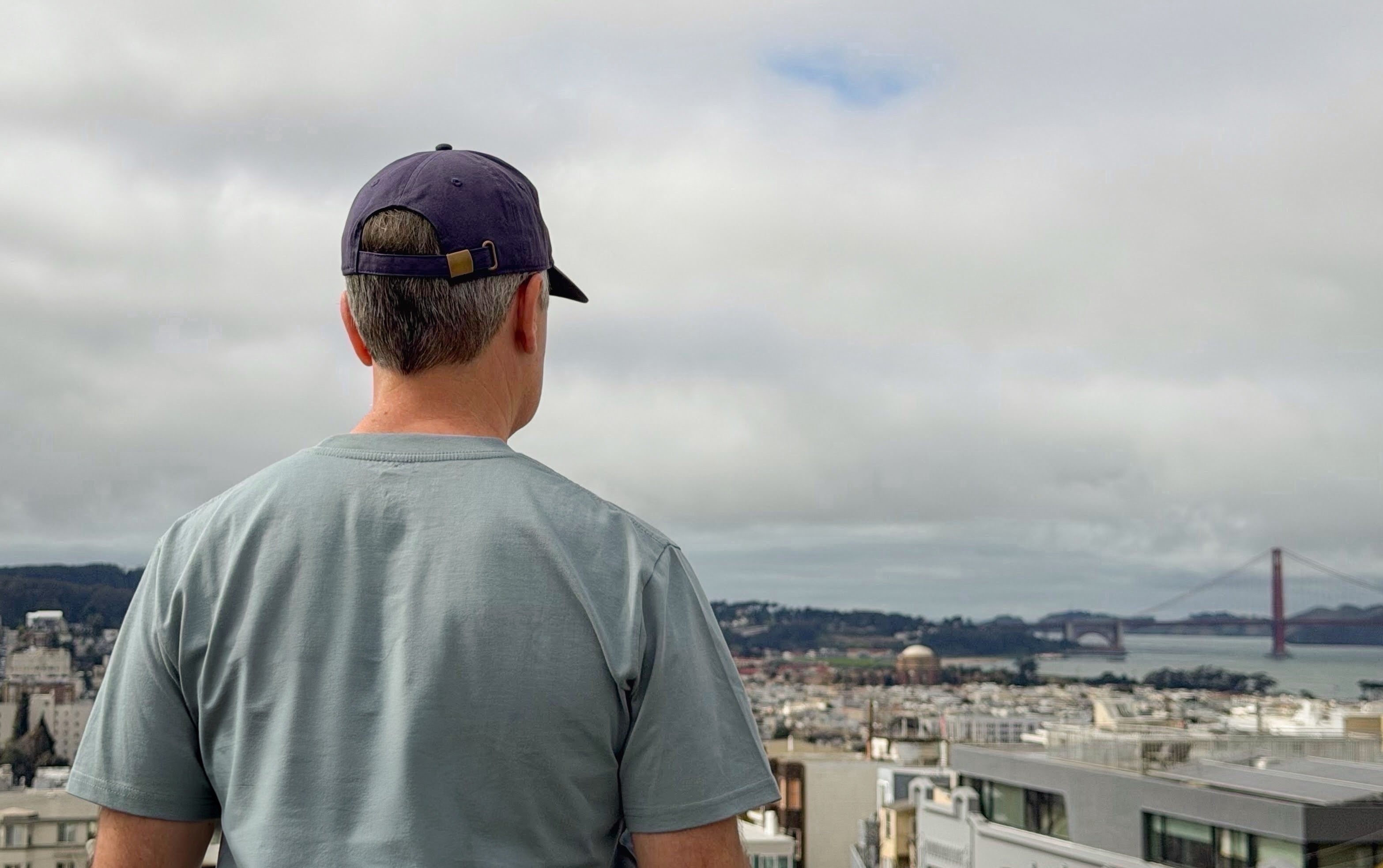 Man wearing an Avelamer Caledonia Cap in Deepwater navy and Ebbtide Tee in Shallow Bay blue-green, looking at the Golden Gate Bridge and the San Francisco cityscape in the distance.