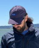 Man wearing a navy cap with a logo and sunglasses, standing outdoors near water.