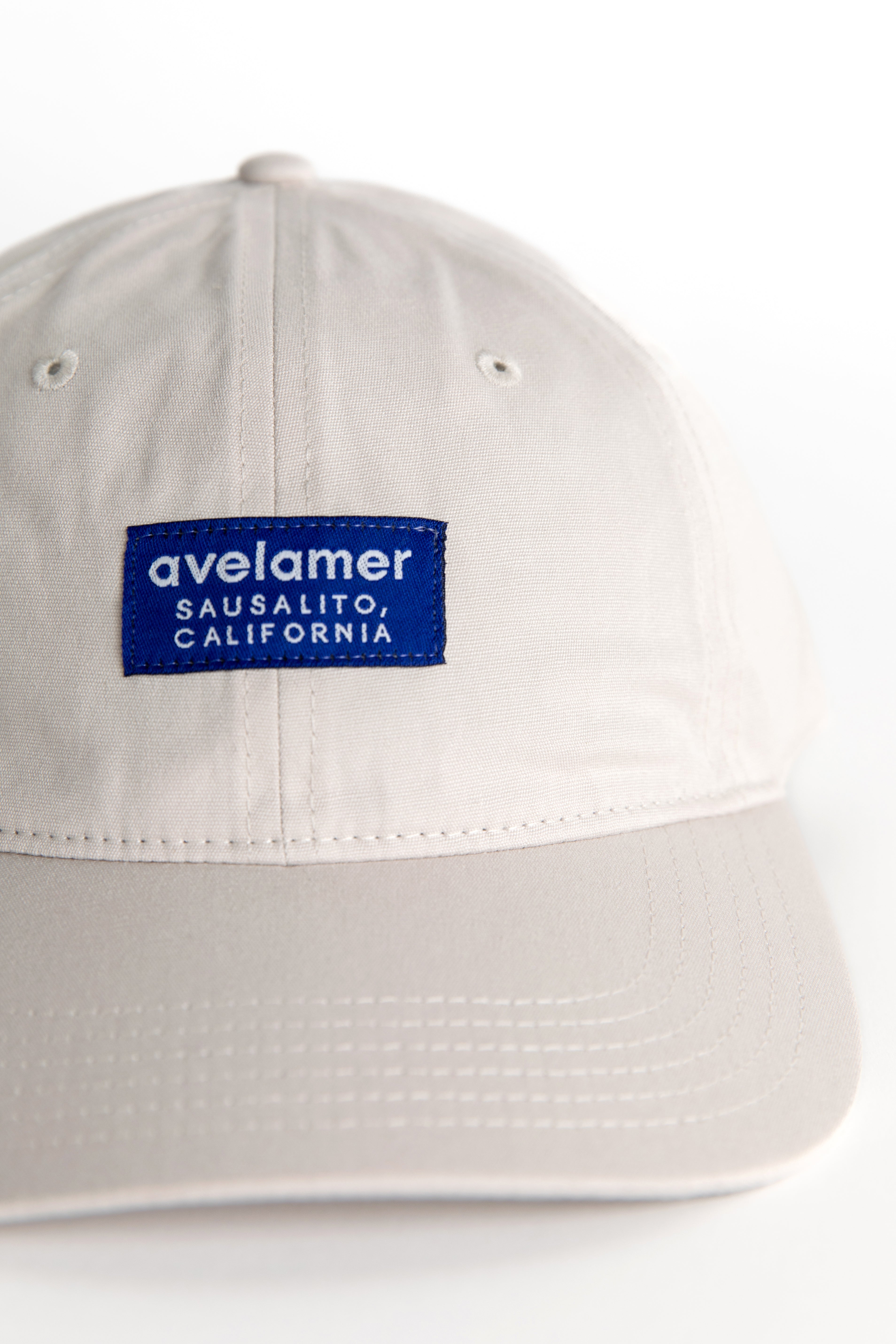 Detail view of an Avelamer Bridgeway Cap in Mainsail (off-white) with a navy Avelamer brand woven label on the front panel