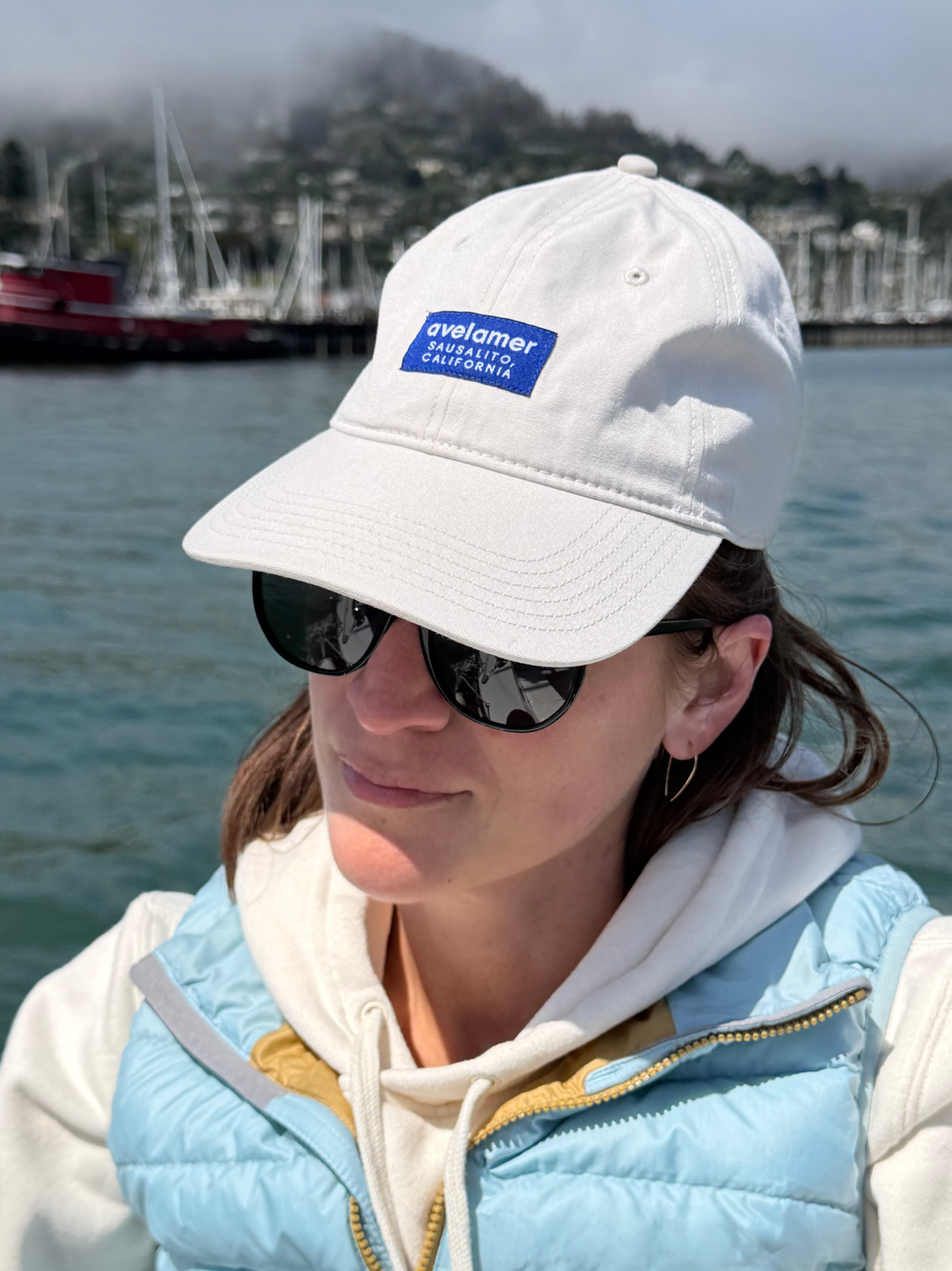 A woman wearing an off-white cap with a navy woven label logo and sunglasses, standing by the Sausalito waterfront.