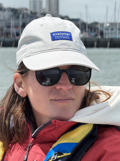 Woman wearing off-white cap with navy label, sunglasses, sailing jacket, with water and bay in the background.