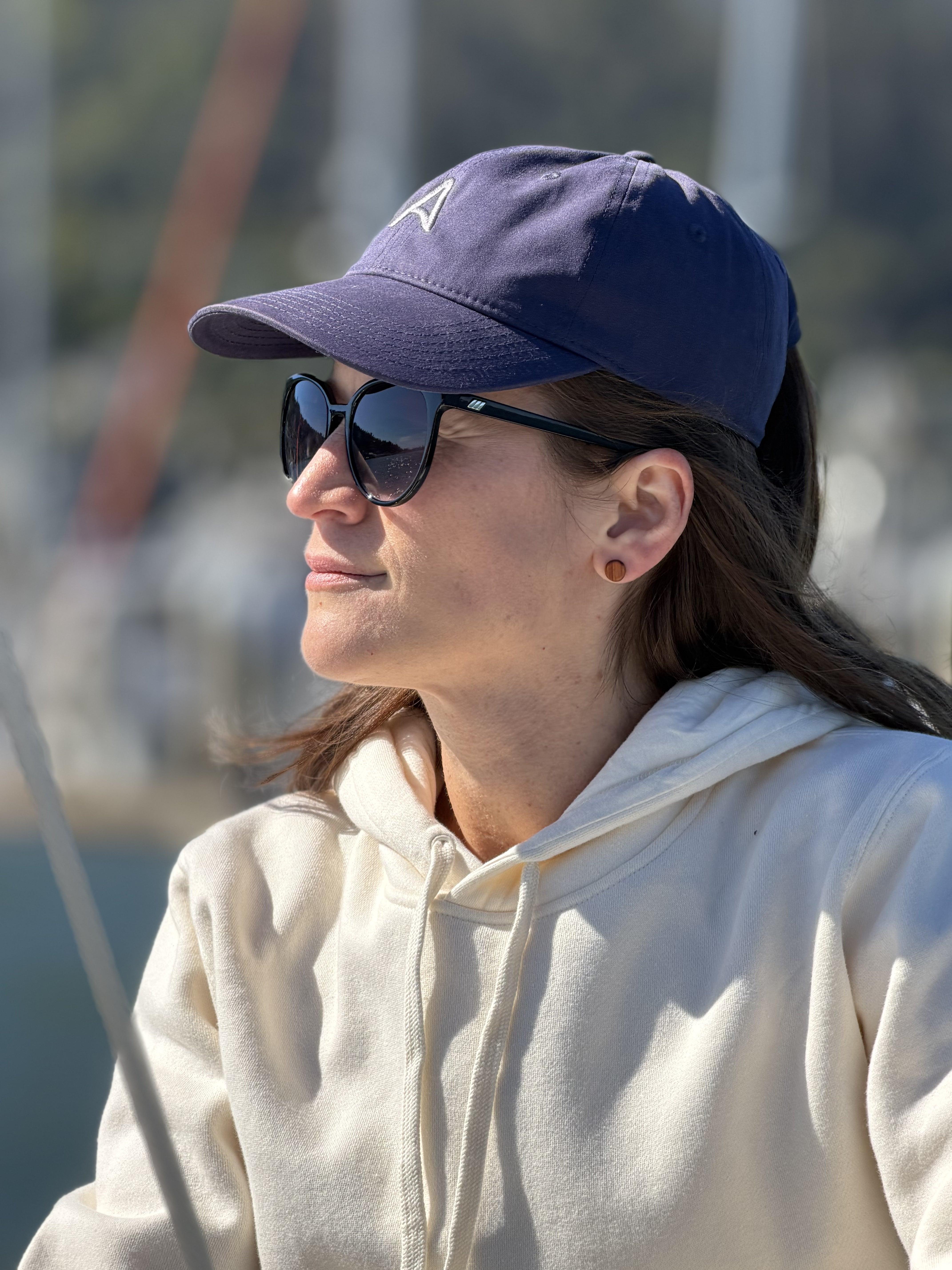 Woman wearing an Avelamer Caledonia Cap in Deepwater navy Sausalito Hoodie in Mainsail off-white with a blurred marina background