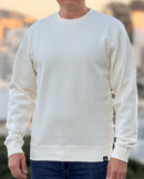 Person wearing an Avelamer Cazneau Crew in Mainsail (off-white) sweatshirt with an Avelamer A-flag logo woven brand hem label visible, against a blurred background