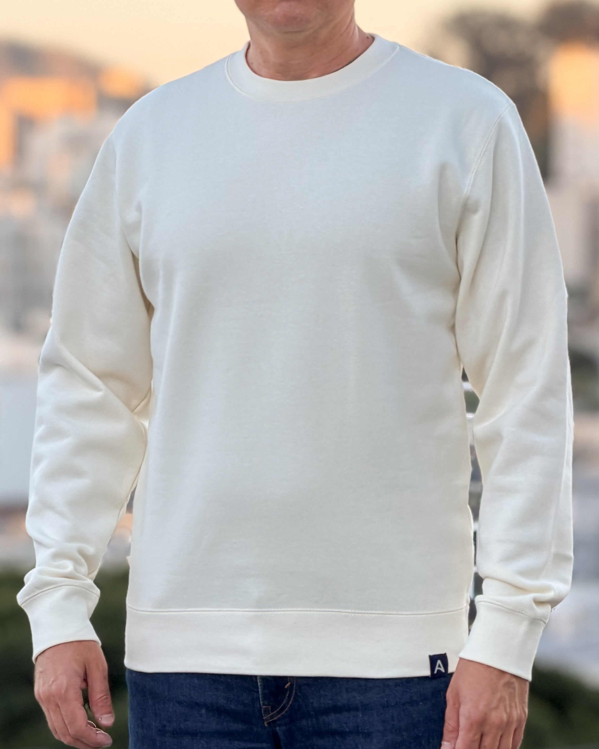 Person wearing an Avelamer Cazneau Crew in Mainsail (off-white) sweatshirt with an Avelamer A-flag logo woven brand hem label visible, against a blurred background