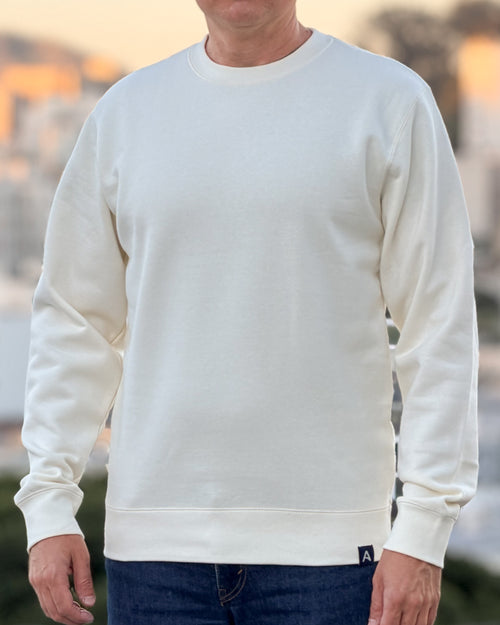 Person wearing an Avelamer Cazneau Crew in Mainsail (off-white) sweatshirt with an Avelamer A-flag logo woven brand hem label visible, against a blurred background