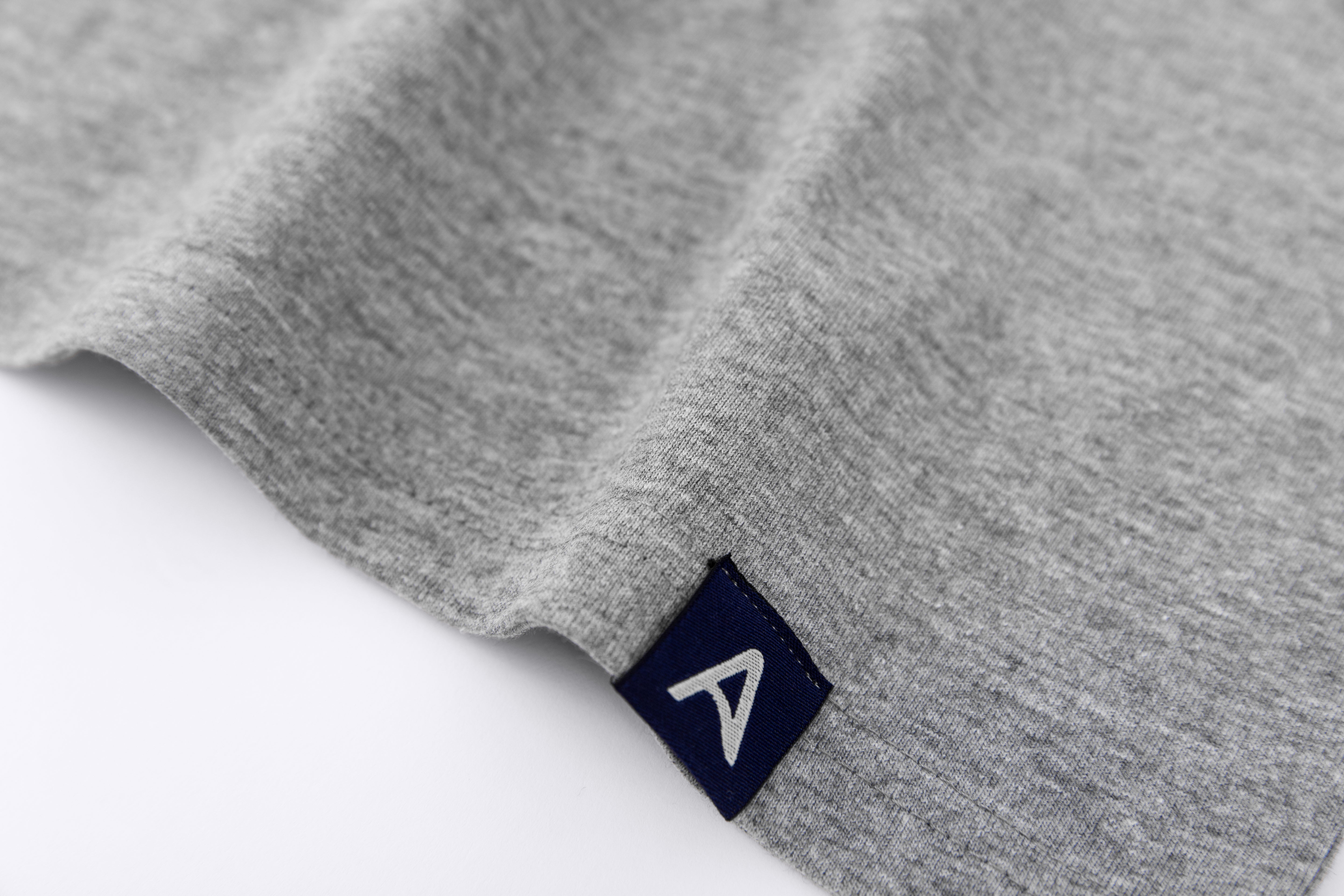 Close-up of Avelamer Ebbide Tee in Morning Fog heather gray with blue A-flag logo woven hem label