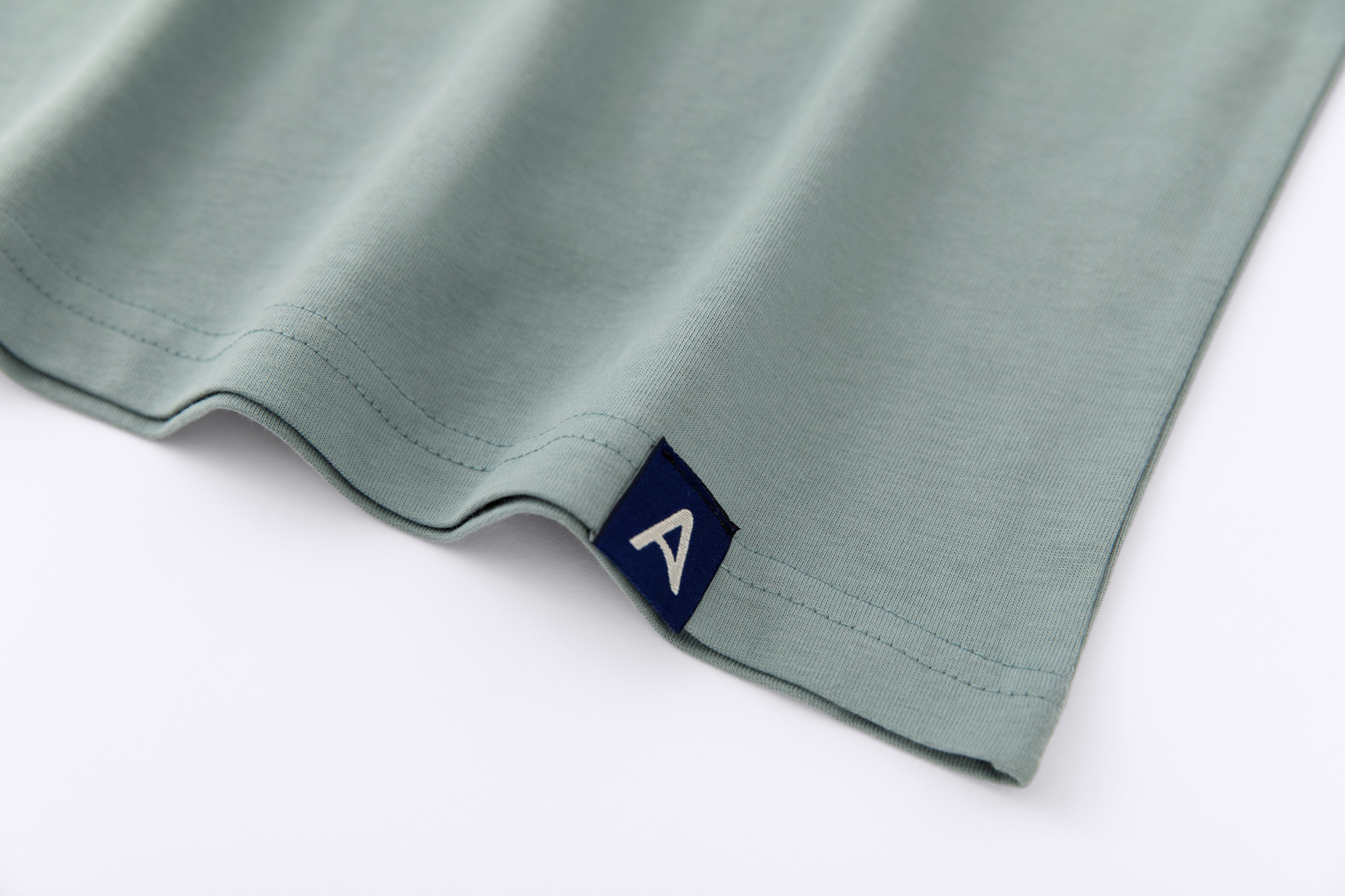 Close-up of Avelamer Ebbide Tee in Shallow Bay blue-green with blue A-flag logo woven hem label