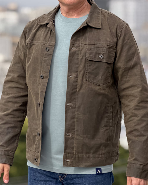 Man wearing an olive jacket over a light blue-green shirt with a blurred background