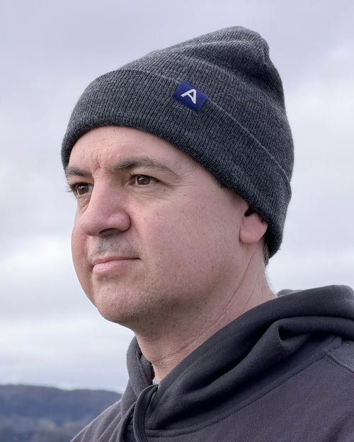 Man wearing an Avelamer Harbor Beanie in Headland Shadow dark gray with a blue woven A-flag label against a cloudy sky