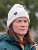 Woman wearing an Avelamer Harbor Beanie in Mainsail off-white with a blue woven A-flag logo label and a green jacket outdoors.