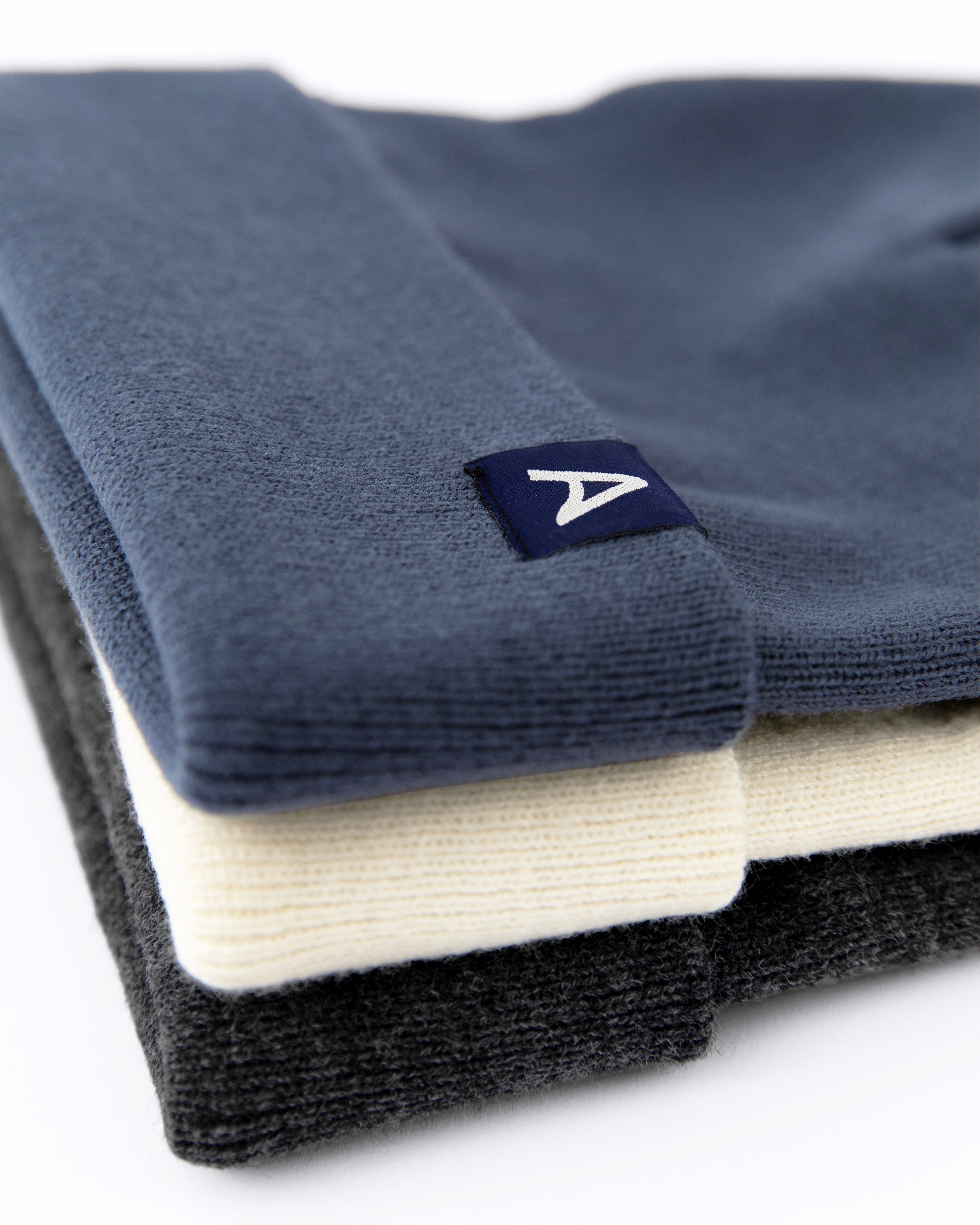 Stack of three Avelamer Harbor Beanies in Ocean Slate blue-gray, Mainsail off-white, and Headland Shadow dark gray with a blue A-flag logo woven cuff label
