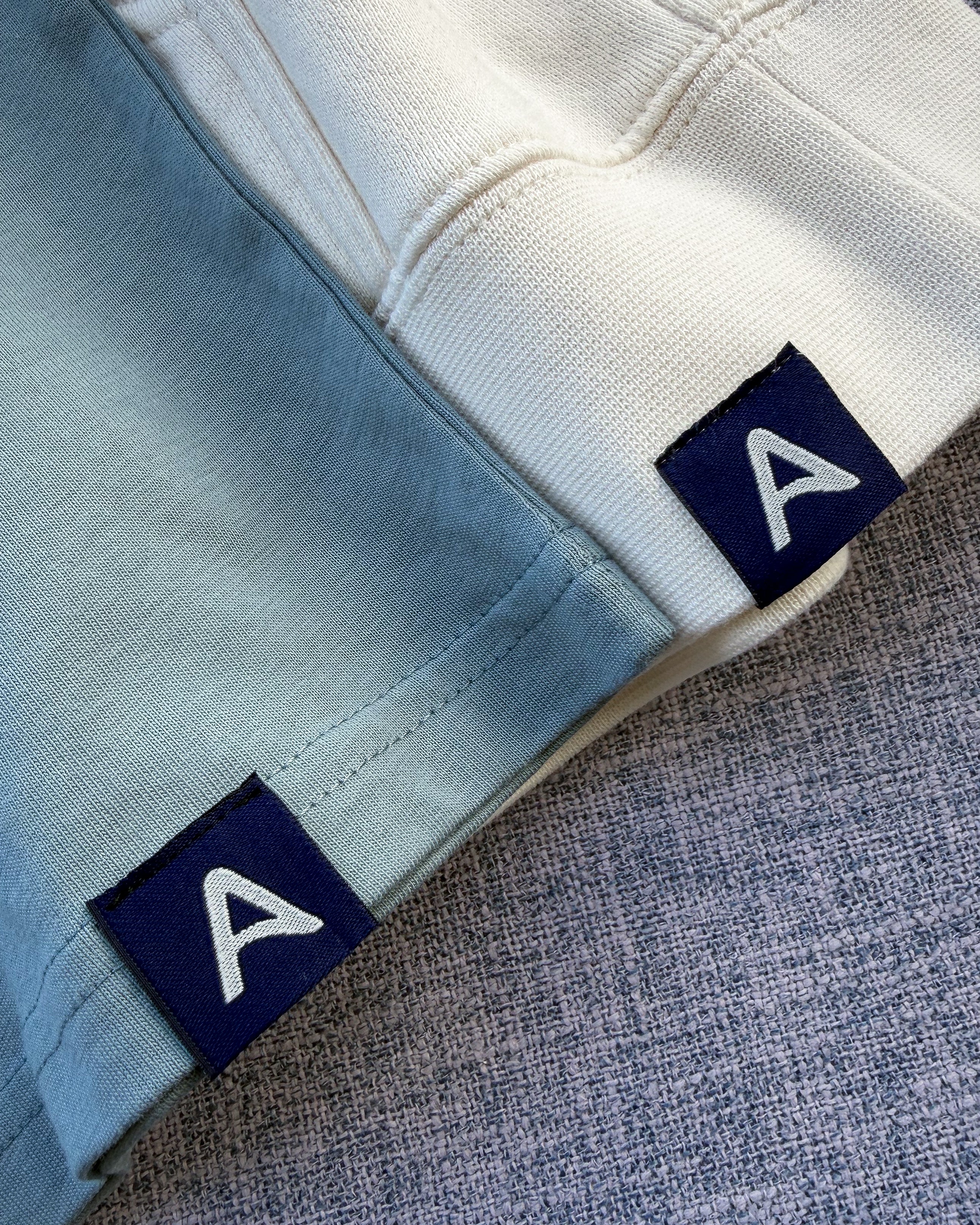 Close-up of Avelamer Sausalito Hoodie in Mainsail (off-white) and Avelamer Ebbite Tee in Shallow Bay (blue-green) with Avelamer A-flag woven hem labels visible. 