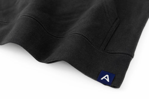 Close-up of Avelamer Sausalito Hoodie in Headland Shadow dark gray with blue A-flag logo woven hem label on a whtie background