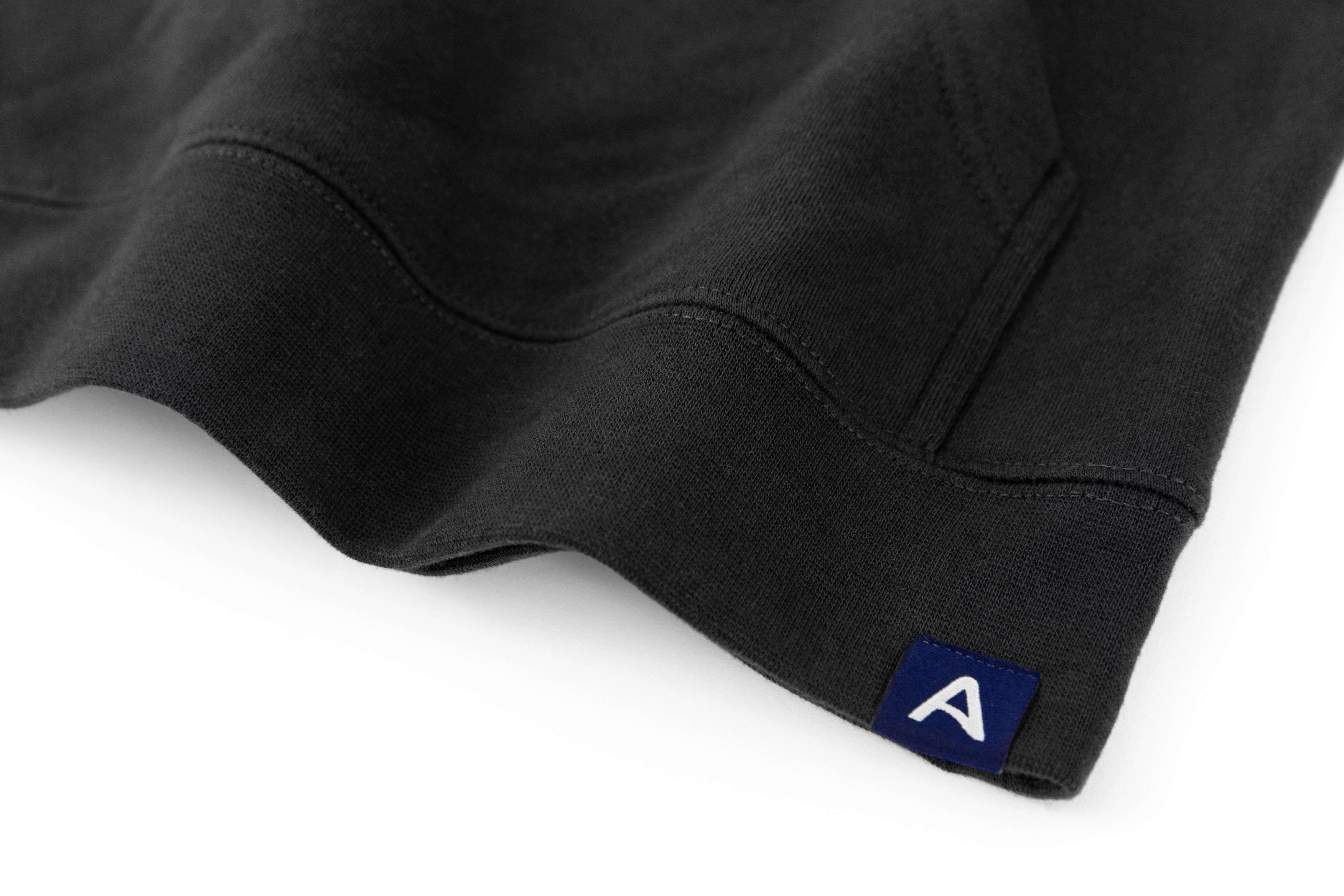 Close-up of Avelamer Sausalito Hoodie in Headland Shadow dark gray with blue A-flag logo woven hem label on a whtie background