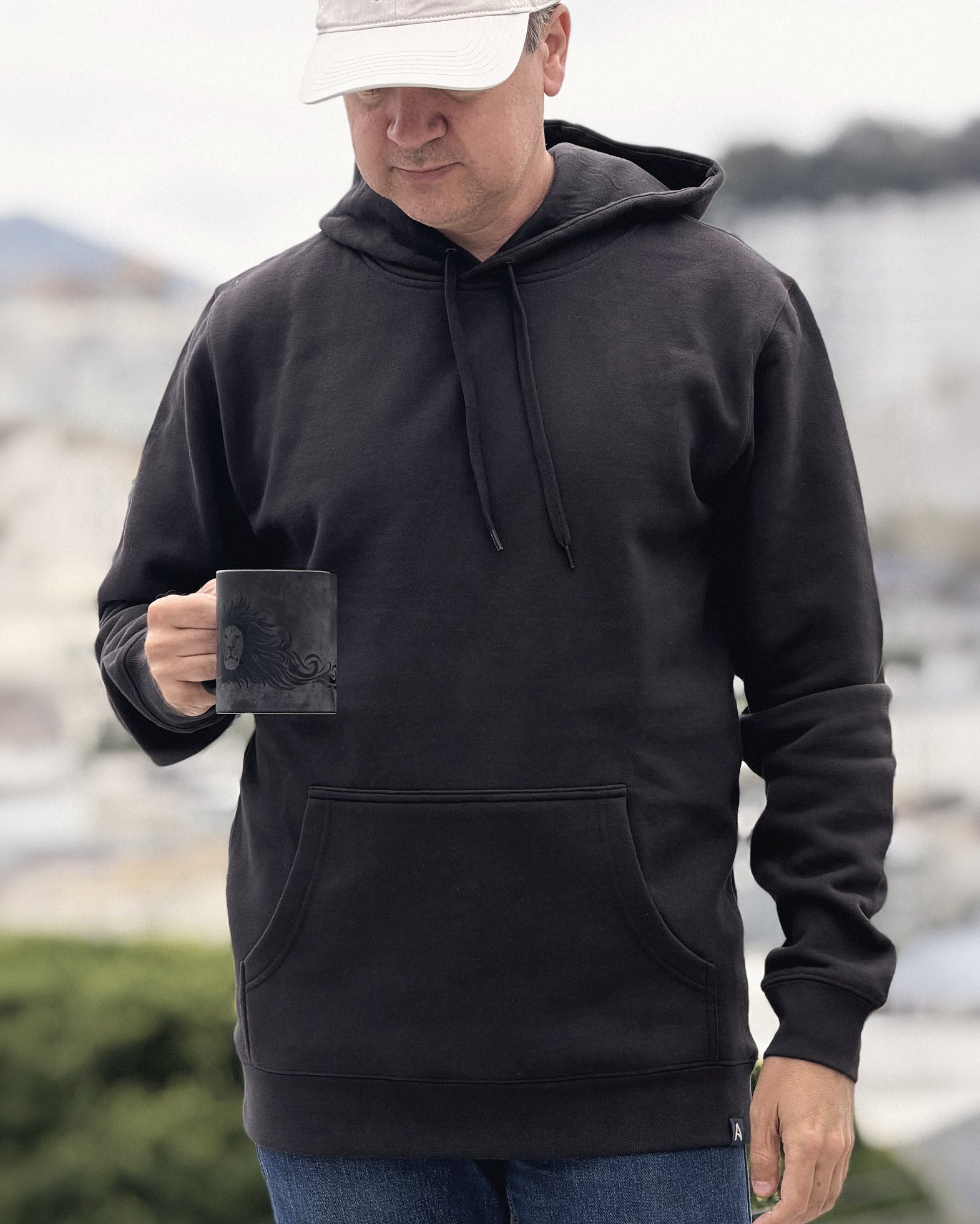 Man wearing the Avelamer Sausalito Hoodie in Headland Shadow, a dark charcoal pullover hoodie with kangaroo pocket, holding a coffee mug outdoors above the San Francisco skyline.