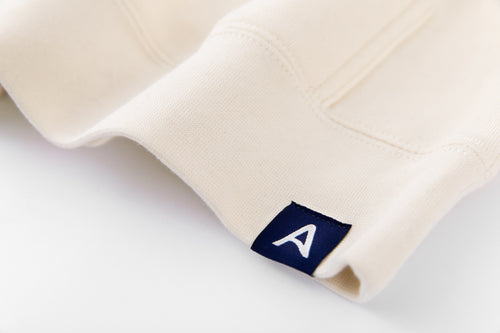 Close-up of Avelamer Sausalito Hoodie in Mainsail off-white with blue A-flag logo woven hem label on a whtie background
