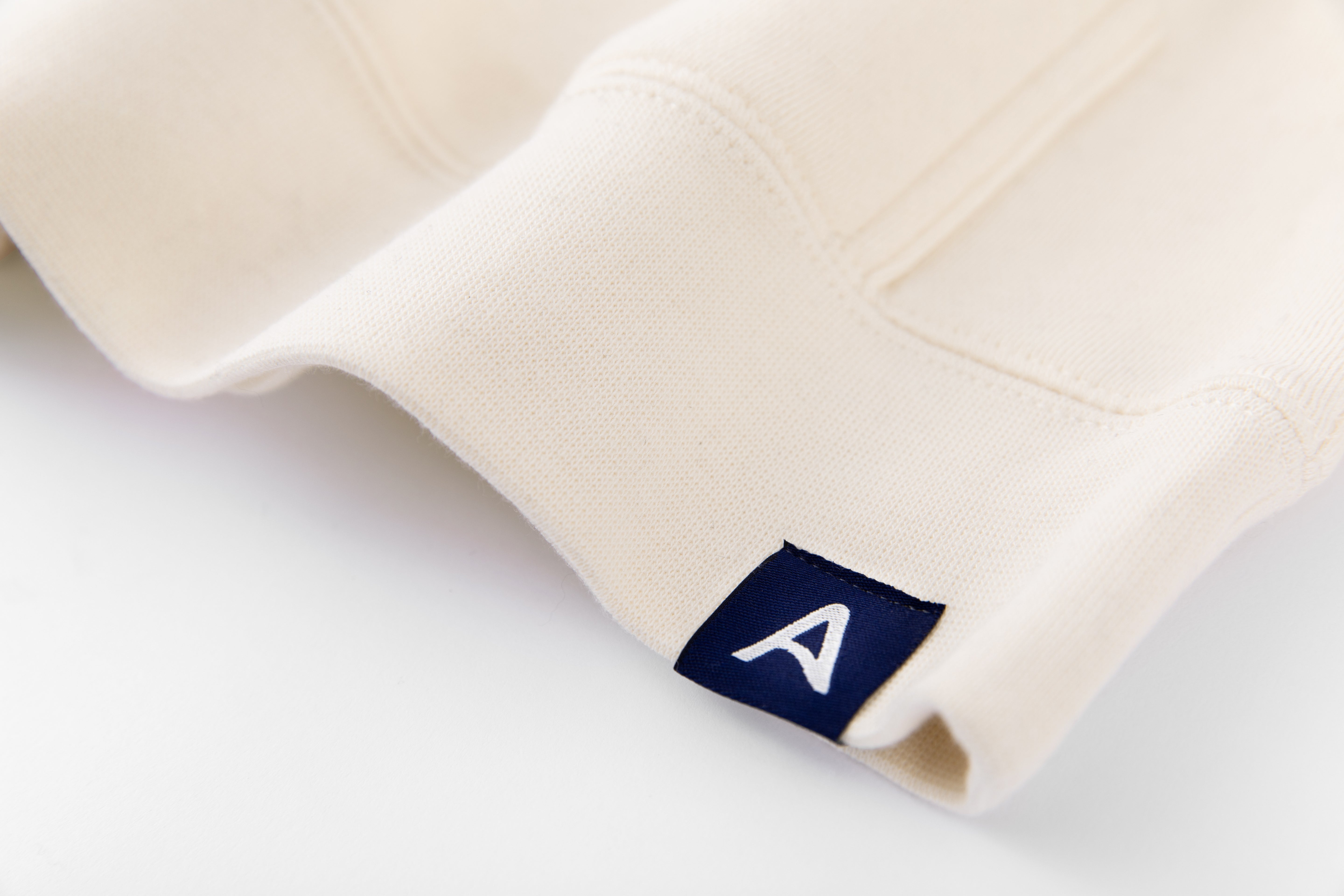 Close-up of Avelamer Sausalito Hoodie in Mainsail off-white with blue A-flag logo woven hem label on a whtie background