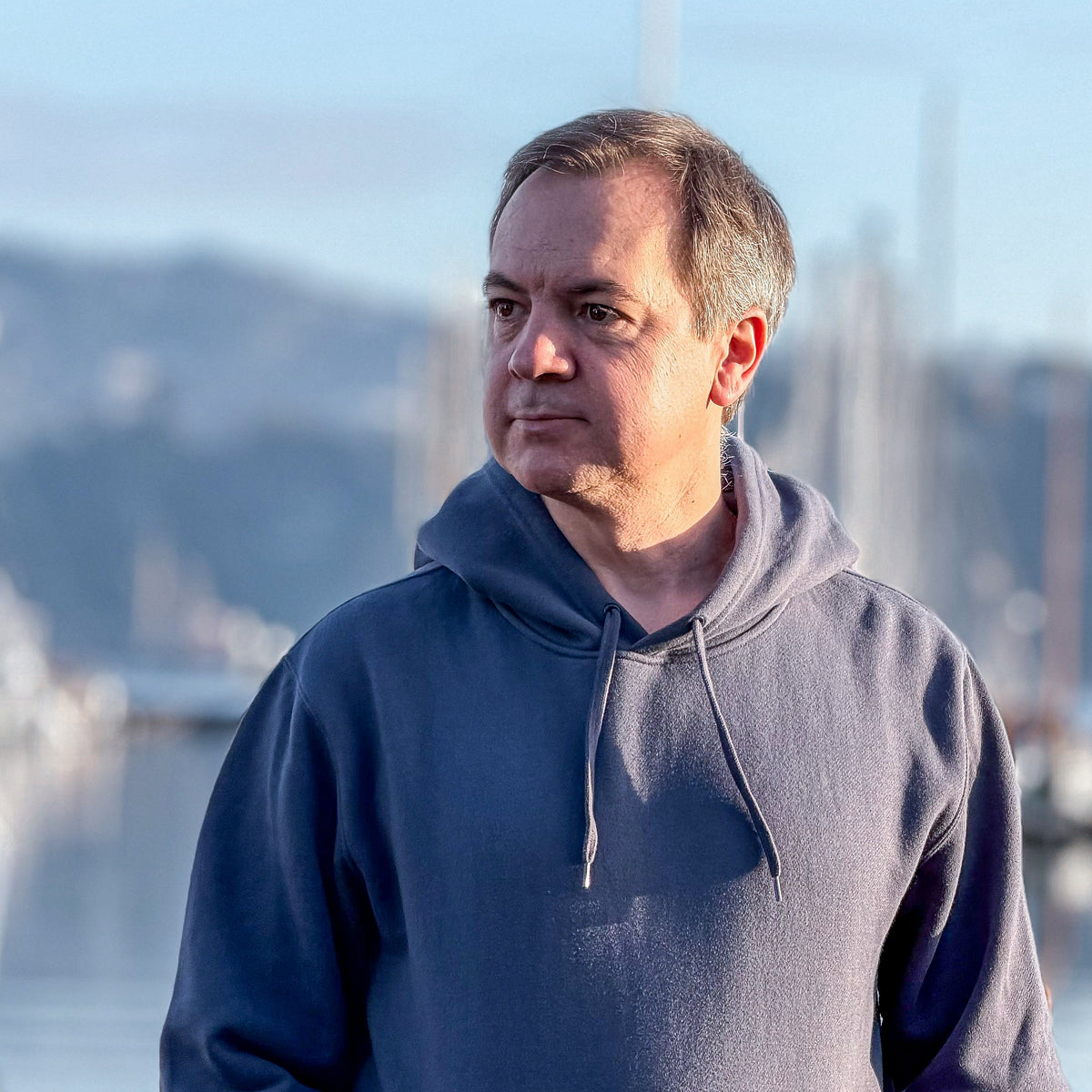 Man wearing an Avelamer Sausalito Hoodie in Ocean Slate (blue gray), standing outdoors at a marina in Sausalito (Norther California) with a blurred background