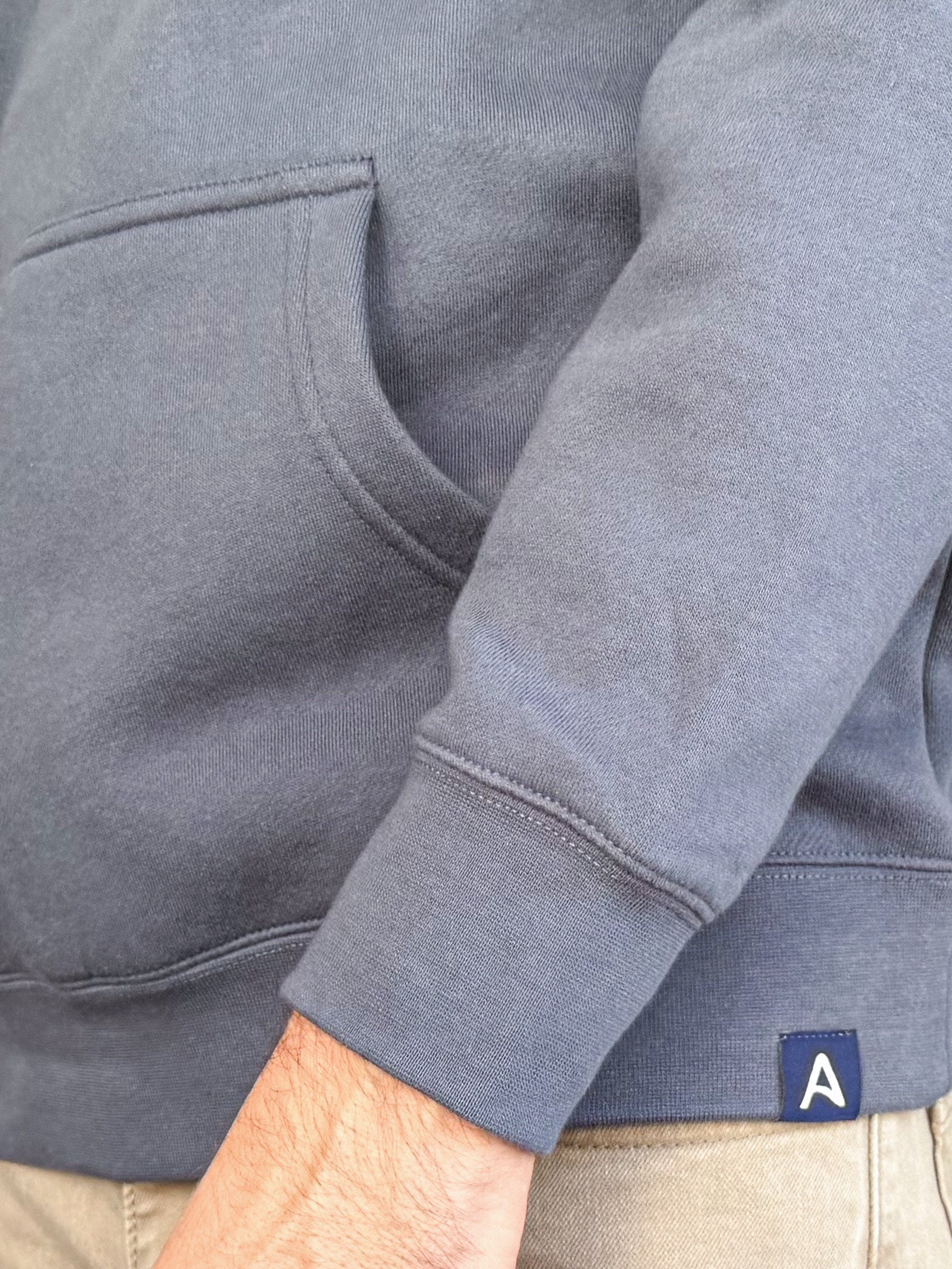Close-up of a person wearing an Avelamer  Sausalito Hoodie in Ocean Slate blue-gray with the blue A-flag logo woven hem label visible. 