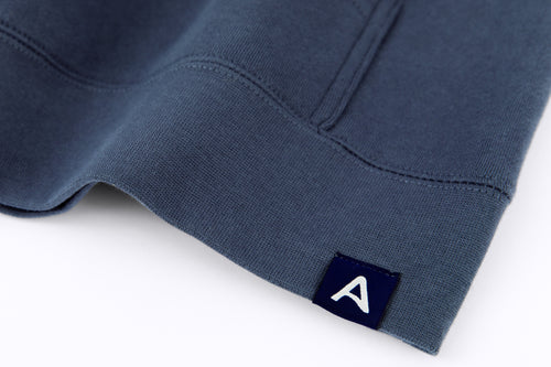 Close-up of Avelamer Sausalito Hoodie in Ocean Slate blue-gray with blue A-flag logo woven hem label on a white background