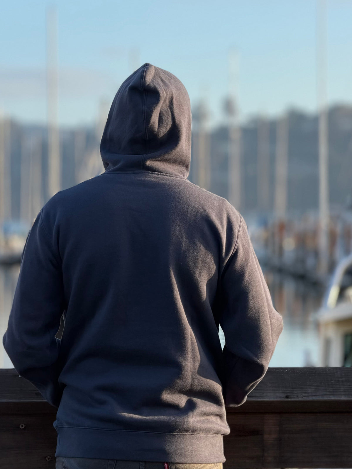 Back view of a person wearing an Avelamer Sausalito Hoodie in Ocean Slate blue-gray looking at a marina