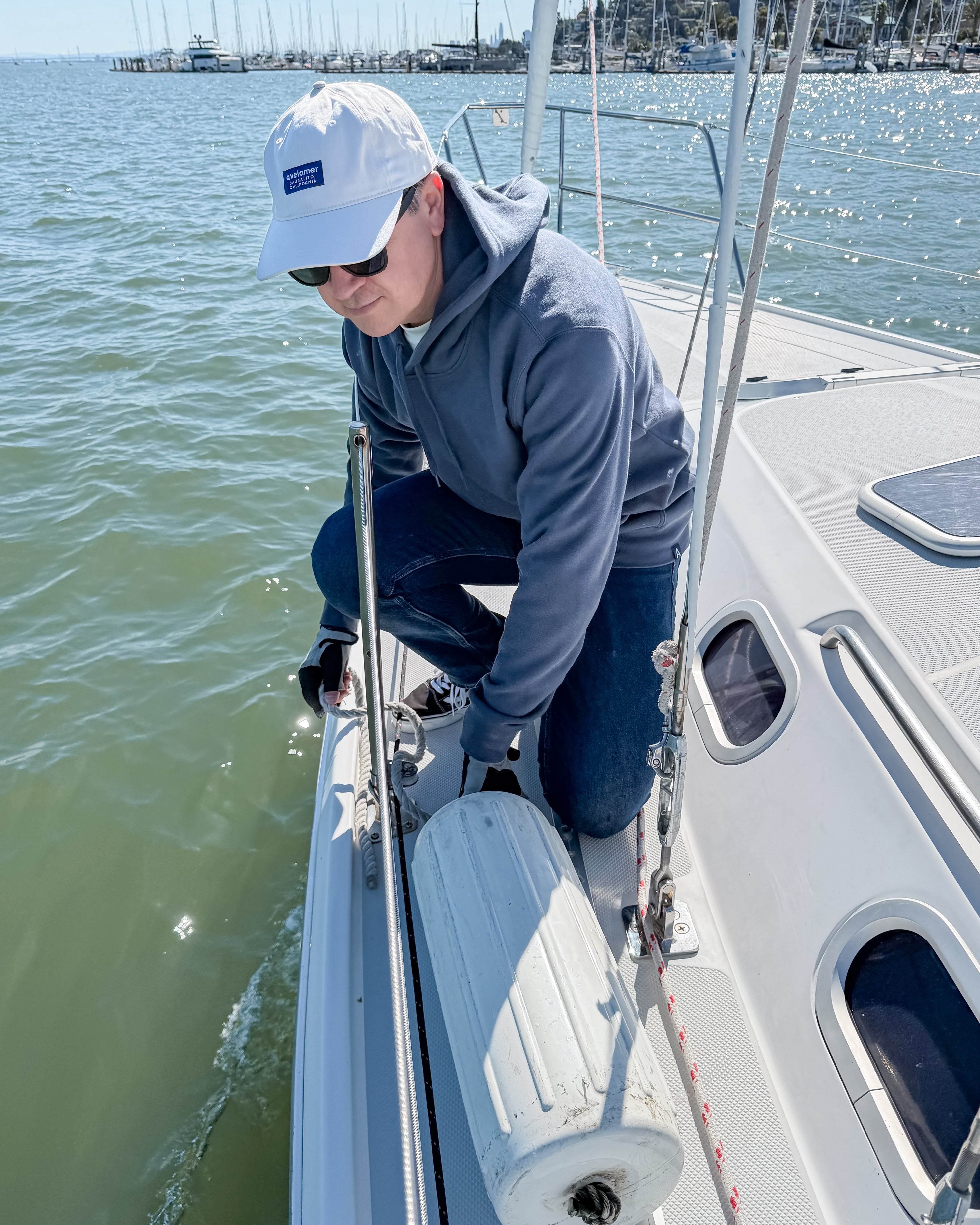 Man on a sailboat in the water in Sausalito, California, wearing an off-white Avelamer cap and blue-gray hoodie.