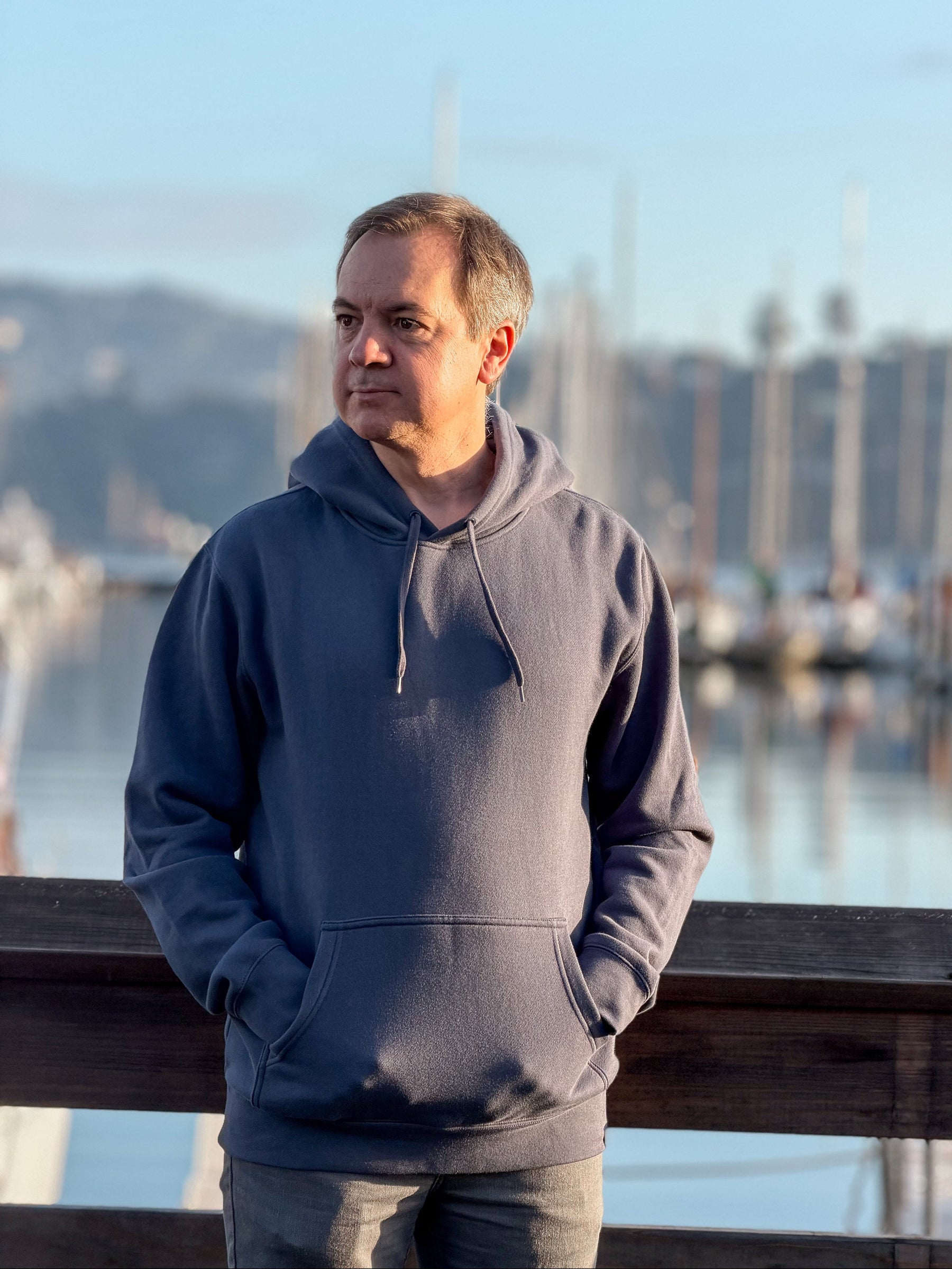 Man wearing an Avelamer Sausalito Hoodie in Ocean Slate blue-gray standing by a marina in Sausalito, California. 