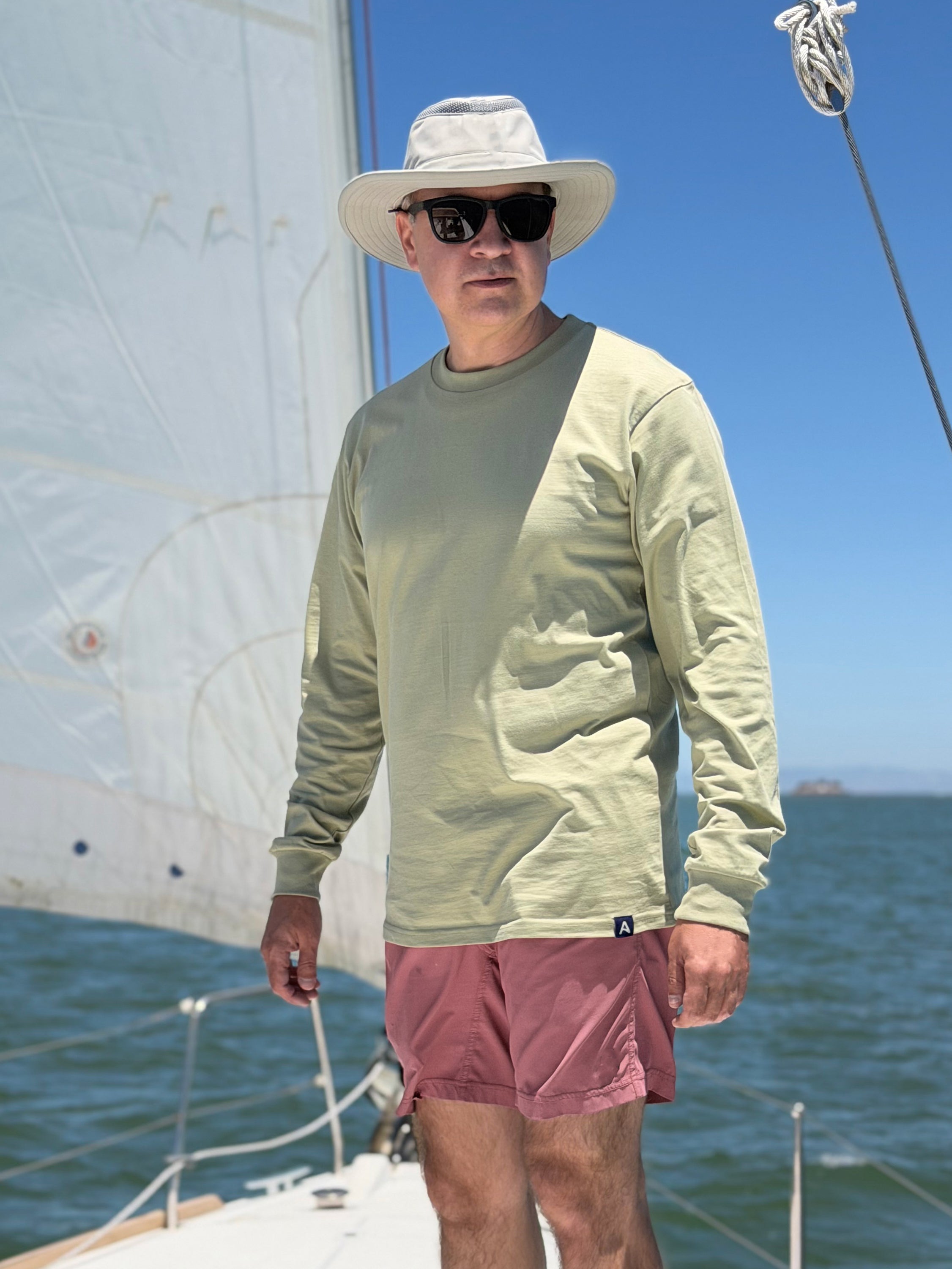 Person wearing an Avelamer Spencer Long Sleeve tee in Bluff Sage (light green) on a sailboat on San Francisco Bay, with an Avelamer A-flag woven hem label.