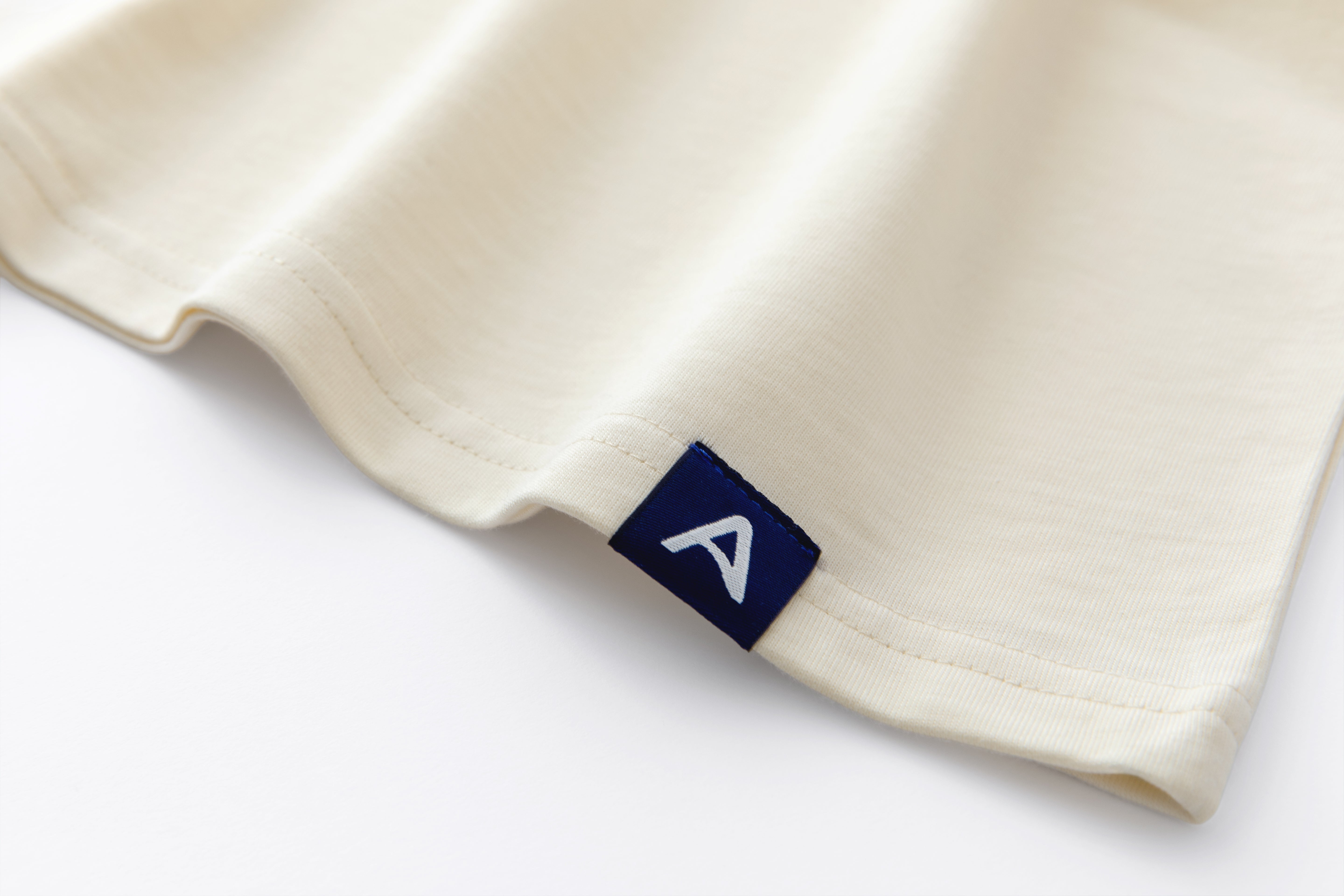 Close-up of Avelamer Spencer Long Sleeve tee in Mainsail off-white with a blue Avelamer A-flag woven hem label. 