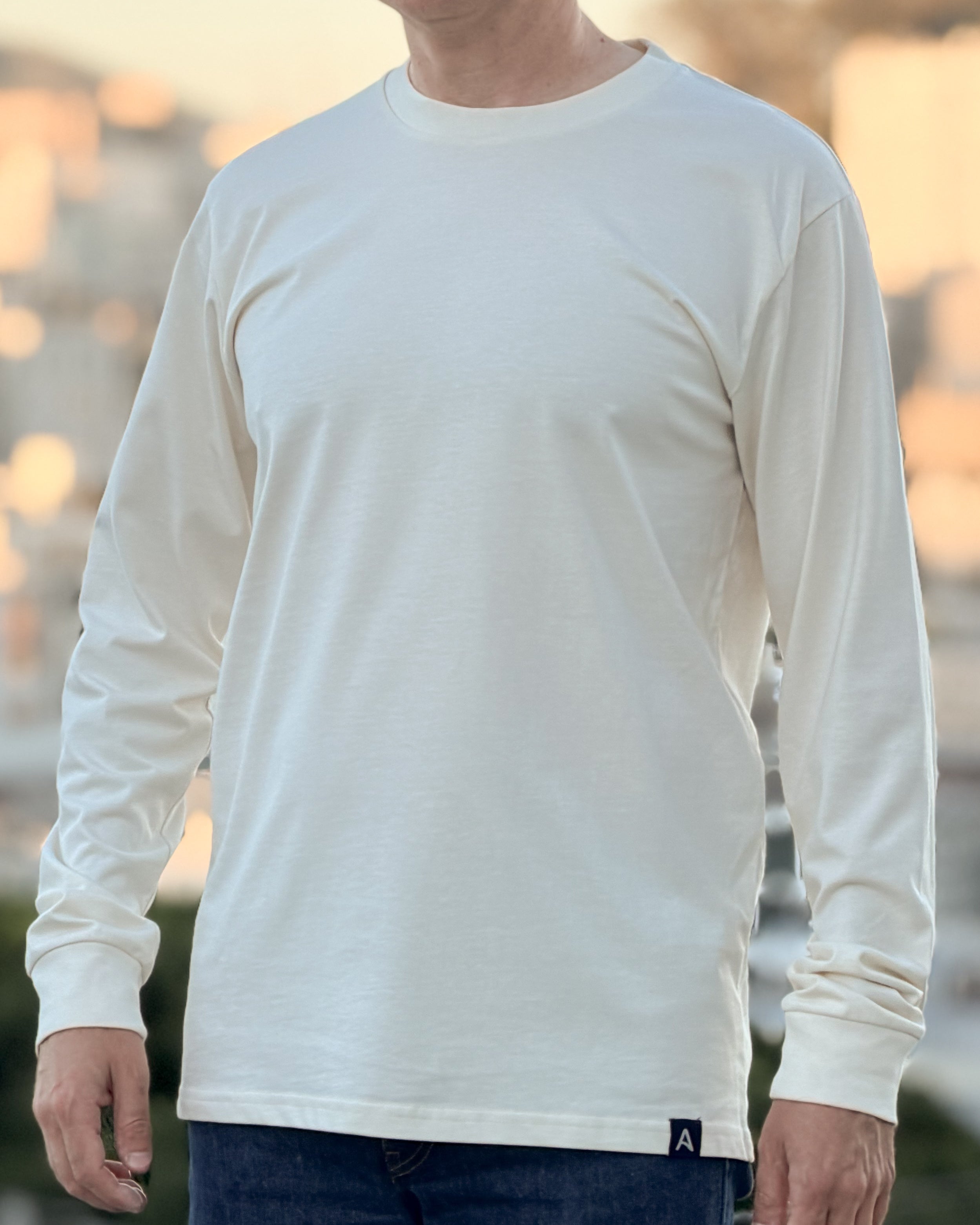 Person wearing an Avelamer Spencer Long Sleeve in Mainsail (off-white) with an Avelamer A-flag hem label, standing against a blurred natural background.