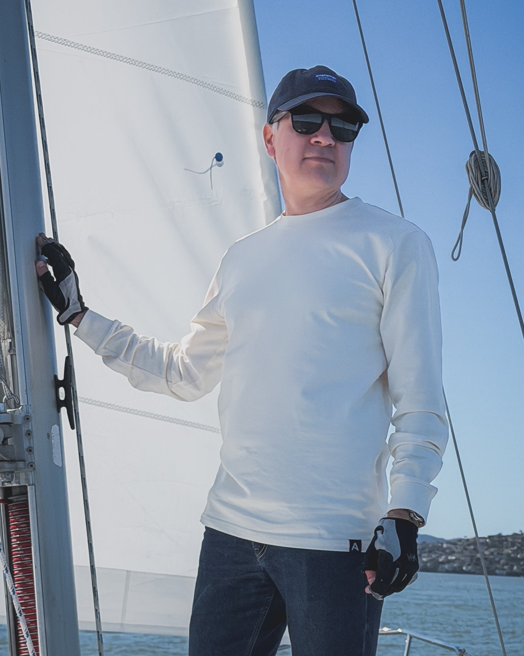 Man on a sailboat wearing an Avelamer Spencer Long Sleeve tee in Mainsail (off-white) and an Avelamer Bridgeway Cap in Deepwater (Navy) with sunglasses, with a clear blue sky background.